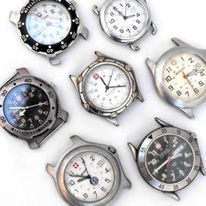May include: A collection of seven stainless steel wristwatches without straps. The watches feature various round dials in black or white, some with rotating bezels, numeric hour markers, and the Wenger or Victorinox logo printed on the watch faces.