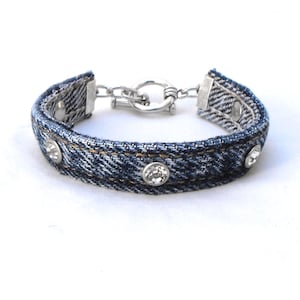 May include: A blue denim bracelet featuring a series of embedded clear round rhinestones along the band. The bracelet is secured with a silver-toned metal toggle clasp, which includes a circular ring and a T-bar closure mechanism.