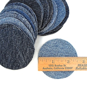 May include: A stack of circular denim fabric patches in various shades of blue, ranging from dark indigo to light wash. A wooden ruler is placed across one light blue denim circle, showing a diameter of approximately 2.5 inches.