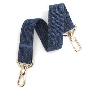 May include: A denim blue bag strap with gold-colored metal clasps. The strap is made of a woven denim fabric and features a darker blue stripe detail. The clasps are a shiny gold color and are designed to attach to a bag.
