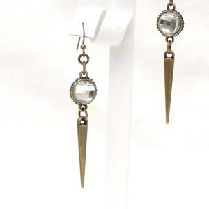 May include: A pair of antique brass-toned earrings. Each earring features a clear, faceted crystal orb, a chain, and a long, pointed spike. The earrings are displayed on a white stand against a white background.