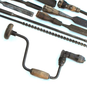May include: A collection of vintage hand tools including a metal brace with a wooden handle, a long twisted metal auger bit, and several wood chisels featuring worn wooden handles and dark, oxidized metal blades arranged on a white surface.