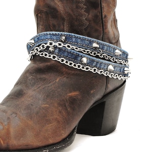May include: A brown leather cowboy boot with a denim and silver chain boot cuff. The cuff features silver spikes and a double chain design, adding a touch of edgy style to the footwear.