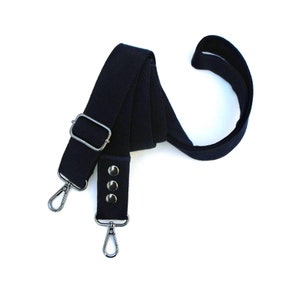 May include: A navy blue adjustable bag strap with silver-tone metal hardware. The strap features a rectangular buckle, three rivets, and two swivel snap hooks. The strap is made of a durable, woven material.