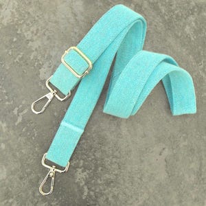 May include: A turquoise bag strap with gold-colored metal hardware. The strap has a woven texture and features two silver-colored swivel clasps and a gold-colored rectangular buckle. The strap is folded over itself in a curved shape.