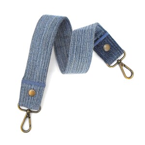 May include: A blue denim bag strap with brass-colored metal clasps and rivets. The strap is made of a woven denim fabric with a light blue and white striped pattern. The ends of the strap are reinforced with solid blue denim.