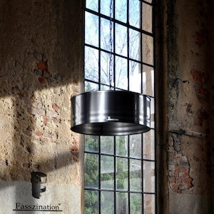 May include: A cylindrical, brushed metal pendant light hangs from a chain in front of a large, multi-paned window. The light fixture is suspended above a rustic wooden table. The background features a weathered brick and plaster wall.