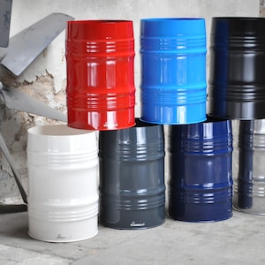 May include: A collection of eight colorful metal barrels in various shades, including red, blue, black, white, gray, and navy. The barrels have a cylindrical shape with horizontal ridges and a shiny finish. The background features an industrial setting with a large fan.
