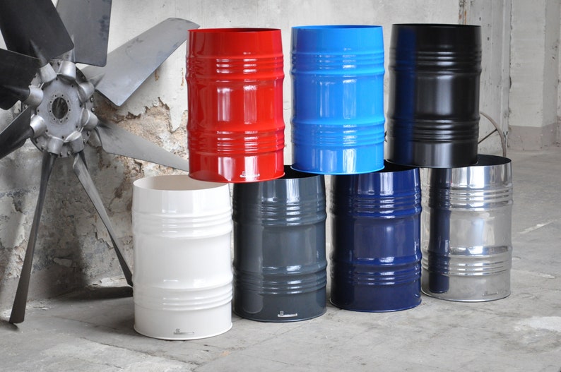 May include: A collection of eight colorful metal barrels in various shades, including red, blue, black, white, gray, and navy. The barrels have a cylindrical shape with horizontal ridges and a shiny finish. The background features an industrial setting with a large fan.