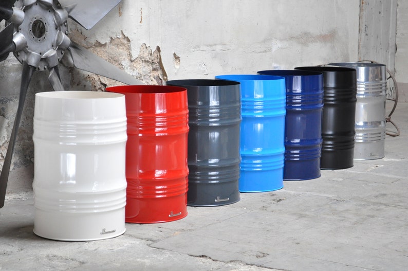 May include: A row of seven colorful metal barrel-shaped containers. The colors include white, red, gray, blue, navy, black, and silver. Each container has horizontal ridges and a smooth, glossy finish. The containers are lined up against a textured wall.