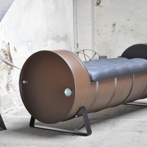 May include: A brown metal barrel repurposed into a bench. The bench has a black cushioned seat and is supported by black metal legs. The barrel's ends are visible, and the overall design has an industrial aesthetic.