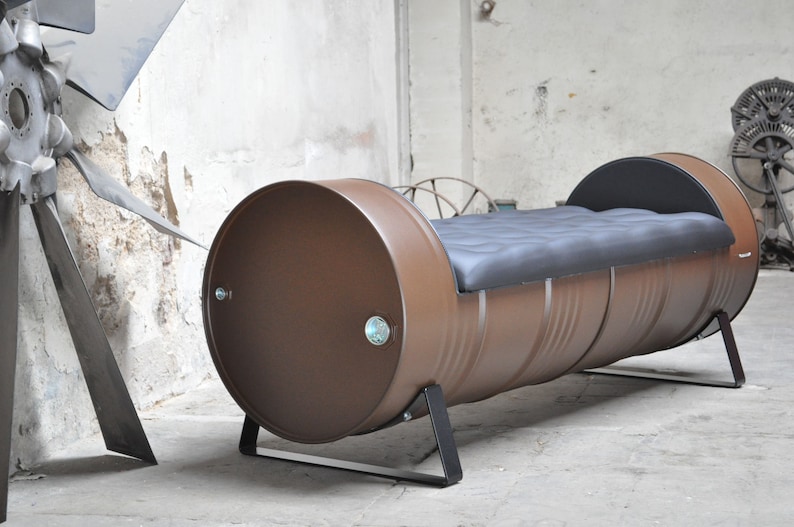 May include: A brown metal barrel repurposed into a bench. The bench has a black cushioned seat and is supported by black metal legs. The barrel's ends are visible, and the overall design has an industrial aesthetic.