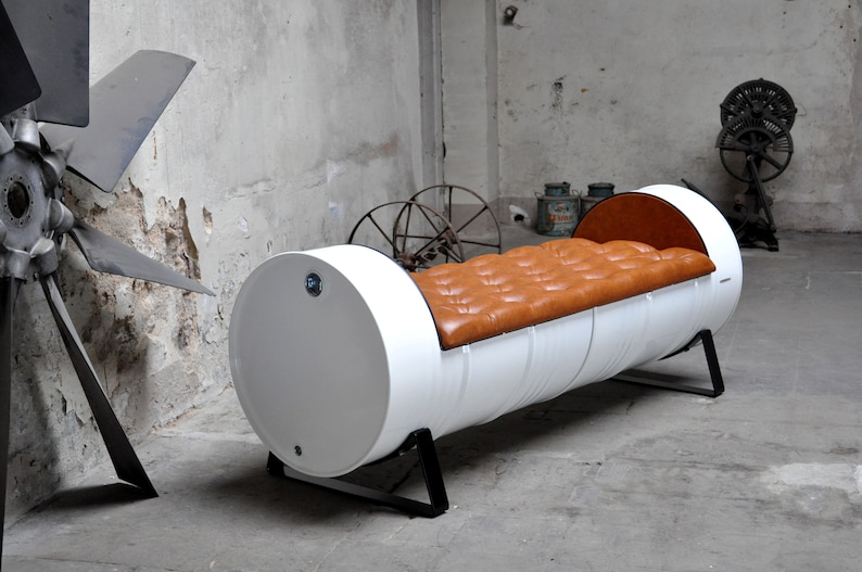 Könnte beinhalten: Weißes, fassförmiges Sofa mit einer gesteppten, braunen Ledersitzfläche. Das Sofa hat schwarze Metallbeine und steht vor einer Betonwand. Das industrielle Design ist ein einzigartiges Möbelstück.