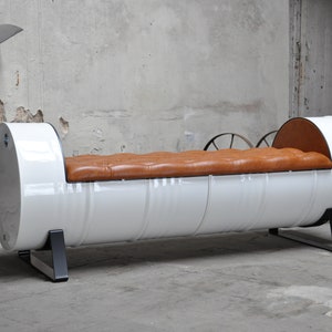 May include: A unique, industrial-style bench made from a repurposed white metal drum. It features a tufted, brown leather seat and black metal legs. The bench is a distinctive piece of furniture.