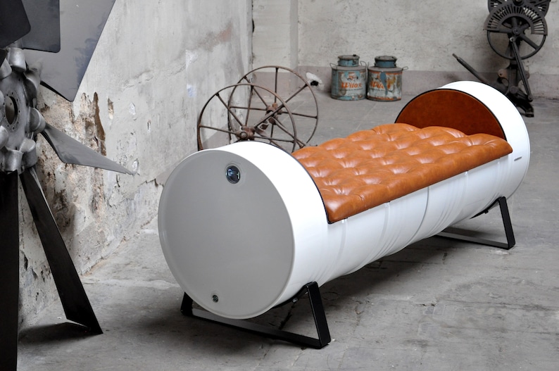 May include: A unique bench made from a repurposed white barrel. The bench features a tufted, brown leather seat and black metal legs. The industrial design is complemented by the barrel's rounded shape and the contrast of colors.