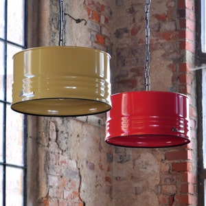 May include: Two industrial-style pendant lights hanging from chains. One is mustard yellow, the other is red. Both have a cylindrical shape with black trim and are suspended against a brick wall background. The lights are designed to resemble oil drums.