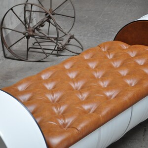 May include: A unique bench made from a repurposed white barrel with a tufted, caramel-colored leather seat. The bench has a curved back and sits on a black metal frame. An antique metal wheel is in the background.
