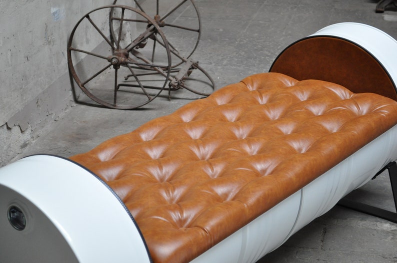 May include: A unique bench made from a repurposed white barrel with a tufted, caramel-colored leather seat. The bench has a curved back and sits on a black metal frame. An antique metal wheel is in the background.
