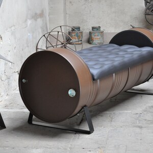May include: A brown metal barrel bench with a black cushioned seat. The bench is supported by black metal legs. The industrial-style furniture piece is set against a concrete wall.