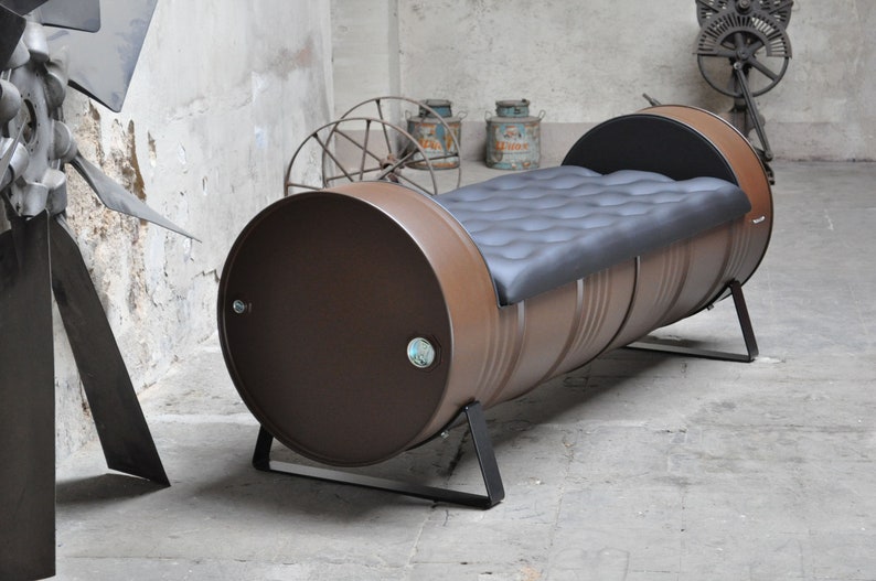 May include: A brown metal barrel bench with a black cushioned seat. The bench is supported by black metal legs. The industrial-style furniture piece is set against a concrete wall.