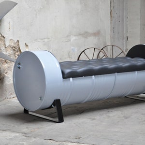 May include: A unique, industrial-style bench made from a repurposed metal drum. The bench is painted light gray with a black, tufted cushion. The bench is supported by black metal legs. The overall design is modern and eco-friendly.