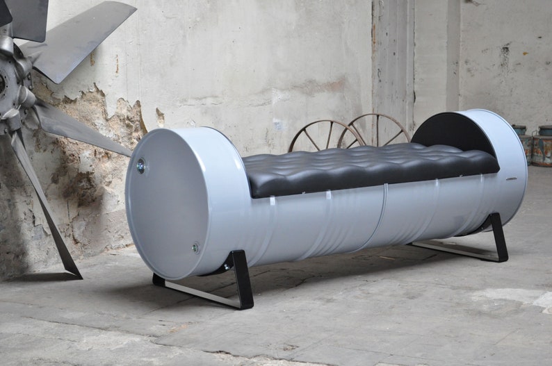 May include: A unique, industrial-style bench made from a repurposed metal drum. The bench is painted light gray with a black, tufted cushion. The bench is supported by black metal legs. The overall design is modern and eco-friendly.