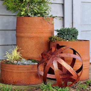 Könnte beinhalten: Eine Ansammlung von rostigen Metallpflanzgefäßen, jeweils mit Grünpflanzen und Sukkulenten gefüllt. Vor den Pflanzgefäßen befindet sich eine dekorative Metallkugel. Die zylindrischen Pflanzgefäße sind unterschiedlich hoch.