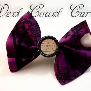 May include: A purple fabric bow with a black and white print of trees and a full moon. The bow has a black button with a white label that reads "AUTHENTIC VAMPIRE FANGS".
