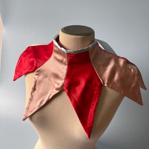 May include: A satin collar featuring alternating pointed panels in vibrant red and soft beige. The neckline is finished with a light blue trim, and the garment is secured with a matching light blue ribbon tie at the back of the neck.