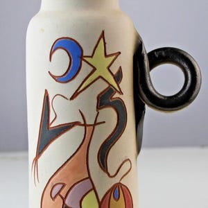 Könnte beinhalten: Cremefarbene Keramikvase mit schwarzem Henkel und Rand. Die Vase ist mit abstrakten Formen in Blau, Gelb, Schwarz, Orange und Rot verziert. Das Design umfasst einen Halbmond, einen Stern und andere geometrische Formen.