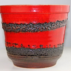 Könnte beinhalten: Ein leuchtend roter Keramik-Blumentopf mit einem strukturierten, dunkelgrauen Band. Der Topf hat eine glänzende Oberfläche und eine breite, abgerundete Form, geeignet für den Innen- oder Außenbereich. Das Design zeigt einen kräftigen Kontrast.