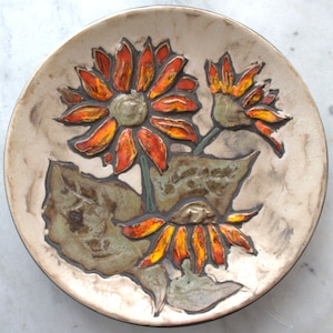 Pode incluir: Um prato de cerâmica com um design em relevo de três girassóis em tons de laranja, amarelo e castanho. O prato tem um fundo branco com uma borda castanha.