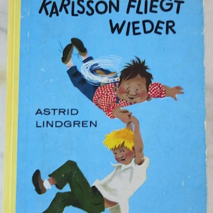 Può includere: Copertina di un libro per bambini vintage con il titolo "KARLSSON FLIEGT WIEDER" in nero su sfondo azzurro. La copertina presenta illustrazioni di due ragazzi a mezz'aria, uno con una camicia a scacchi rossi e bianchi. È visibile anche il nome dell'autore, Astrid Lindgren.