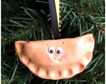 May include: A tan, crescent-shaped pierogi ornament with a crimped edge, googly eyes, and a small smile. The ornament hangs from a black and yellow ribbon and features the text Pittsburgh Pete printed in white below the object.