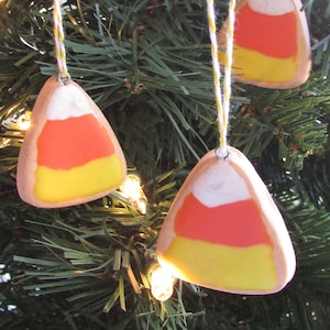 Puede incluir: Tres adornos de ma&iacute;z dulce hechos a mano colgando de un &aacute;rbol de Navidad verde. Los adornos est&aacute;n hechos de arcilla polim&eacute;rica y est&aacute;n pintados en los colores tradicionales del ma&iacute;z dulce, naranja, amarillo y blanco.