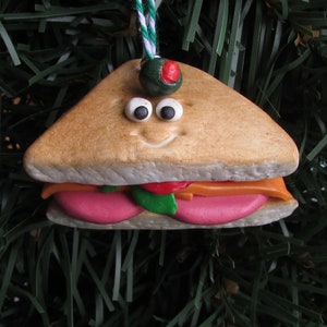 May include: A handmade Christmas ornament shaped like a sandwich with a smiling face. The sandwich is made of brown bread, pink ham, orange cheese, and green lettuce. The ornament has a green olive on top.