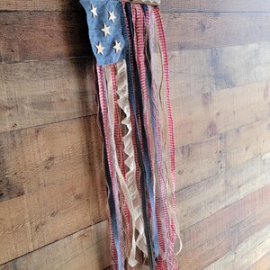 K&ouml;nnte beinhalten: Eine patriotische Wanddekoration, die die amerikanische Flagge darstellt. Sie hat ein blaues Denim-Quadrat mit Holzsternen und roten, wei&szlig;en und blauen B&auml;ndern. Die Oberseite ist mit wei&szlig;er Spitze verziert und h&auml;ngt an einem Naturfaden.