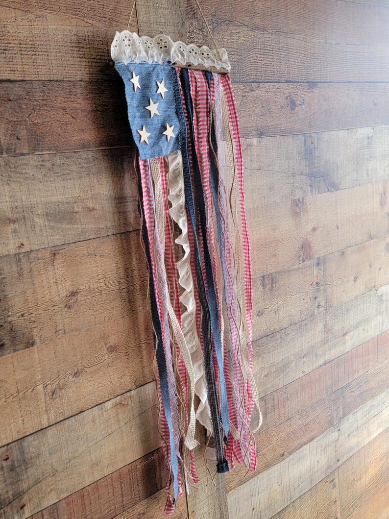 K&ouml;nnte beinhalten: Eine patriotische Wanddekoration, die die amerikanische Flagge darstellt. Sie hat ein blaues Denim-Quadrat mit Holzsternen und roten, wei&szlig;en und blauen B&auml;ndern. Die Oberseite ist mit wei&szlig;er Spitze verziert und h&auml;ngt an einem Naturfaden.
