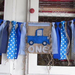 May include: A fabric banner featuring a central burlap pennant with a blue truck applique and the word ONE in grey felt letters. The banner is decorated with various blue and white patterned fabric strips including polka dots and gingham checks.