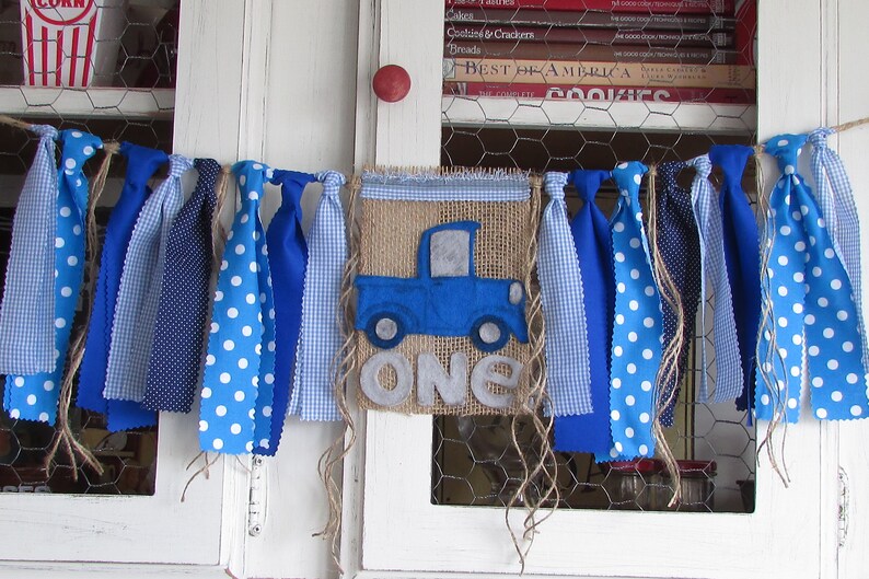 May include: A fabric banner featuring a central burlap pennant with a blue truck applique and the word ONE in grey felt letters. The banner is decorated with various blue and white patterned fabric strips including polka dots and gingham checks.