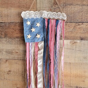 K&ouml;nnte beinhalten: Eine patriotische Wanddekoration, die die amerikanische Flagge darstellt. Sie hat ein denimblaues Quadrat mit Holzsternen, eine wei&szlig;e Spitzentrimmung und rot-wei&szlig; gestreifte B&auml;nder. Die B&auml;nder h&auml;ngen vertikal und bilden ein dekoratives St&uuml;ck.