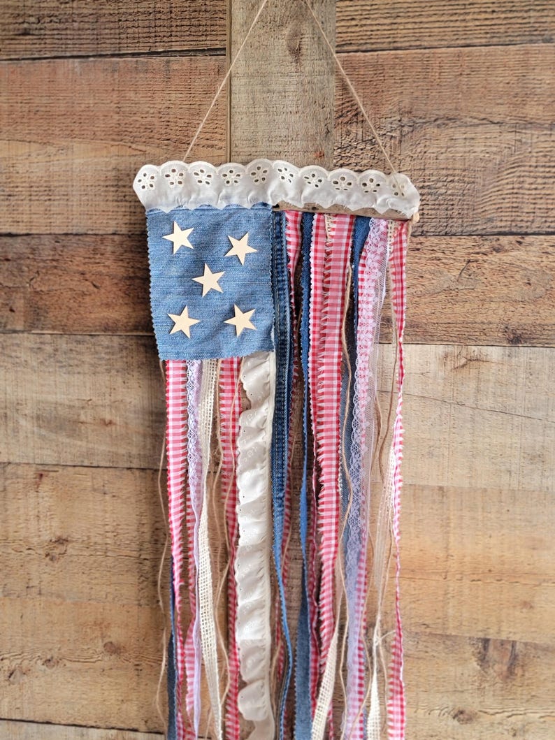 K&ouml;nnte beinhalten: Eine patriotische Wanddekoration, die die amerikanische Flagge darstellt. Sie hat ein denimblaues Quadrat mit Holzsternen, eine wei&szlig;e Spitzentrimmung und rot-wei&szlig; gestreifte B&auml;nder. Die B&auml;nder h&auml;ngen vertikal und bilden ein dekoratives St&uuml;ck.