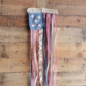K&ouml;nnte beinhalten: Eine patriotische Wanddekoration, die die amerikanische Flagge darstellt. Sie hat ein blaues Jeansquadrat mit Holzsternen und roten, wei&szlig;en und blauen Stoffstreifen. Der obere Rand ist mit wei&szlig;er Spitze verziert und h&auml;ngt an einer Schnur.