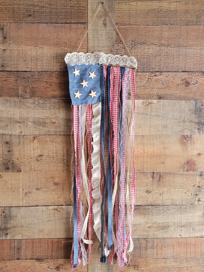K&ouml;nnte beinhalten: Eine patriotische Wanddekoration, die die amerikanische Flagge darstellt. Sie hat ein blaues Jeansquadrat mit Holzsternen und roten, wei&szlig;en und blauen Stoffstreifen. Der obere Rand ist mit wei&szlig;er Spitze verziert und h&auml;ngt an einer Schnur.