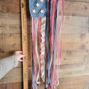 K&ouml;nnte beinhalten: Eine patriotische Wanddekoration, die die amerikanische Flagge darstellt. Sie hat ein denimblaues Quadrat mit Holzsternen, rot-wei&szlig;em Vichy-Karo und verschiedenen B&auml;ndern. Die Oberseite hat eine wei&szlig;e Spitzentrimmung.