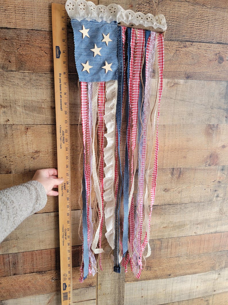 K&ouml;nnte beinhalten: Eine patriotische Wanddekoration, die die amerikanische Flagge darstellt. Sie hat ein denimblaues Quadrat mit Holzsternen, rot-wei&szlig;em Vichy-Karo und verschiedenen B&auml;ndern. Die Oberseite hat eine wei&szlig;e Spitzentrimmung.