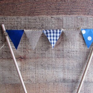 May include: A cake topper featuring five triangular fabric pennants strung between two wooden sticks. The pennants alternate between solid blue, textured beige burlap, blue and white gingham check, and blue with white polka dots.