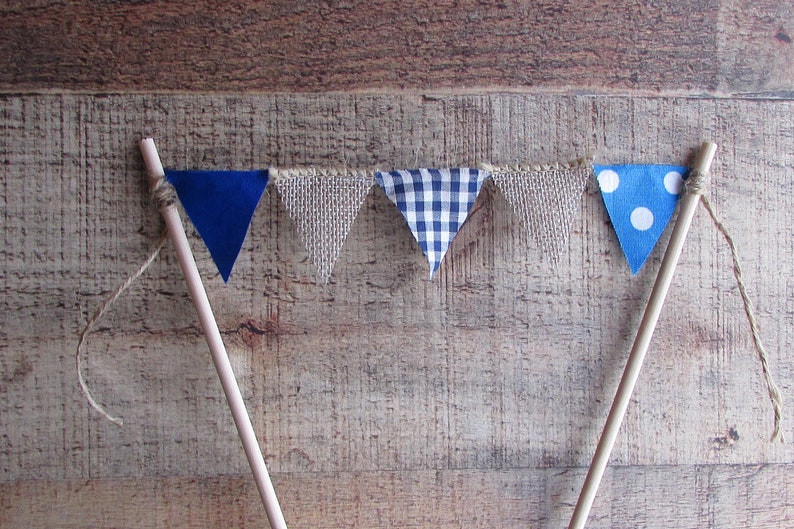 May include: A cake topper featuring five triangular fabric pennants strung between two wooden sticks. The pennants alternate between solid blue, textured beige burlap, blue and white gingham check, and blue with white polka dots.