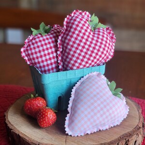 Pu&ograve; includere: Una natura morta rustica presenta una fetta di legno con un cestino di bacche turchese contenente due fragole di stoffa a forma di cuore a quadretti vichy. Una terza fragola di stoffa e due vere fragole completano la composizione. Le fragole di stoffa hanno accenti di foglie verdi.