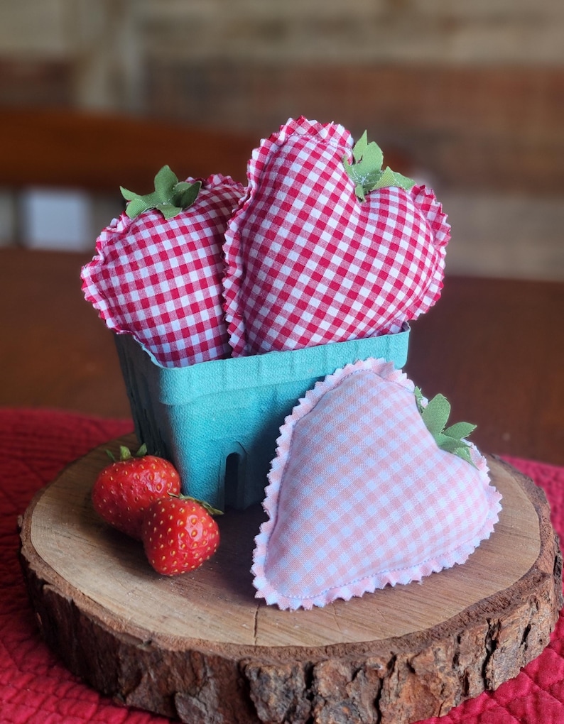 Pu&ograve; includere: Una natura morta rustica presenta una fetta di legno con un cestino di bacche turchese contenente due fragole di stoffa a forma di cuore a quadretti vichy. Una terza fragola di stoffa e due vere fragole completano la composizione. Le fragole di stoffa hanno accenti di foglie verdi.